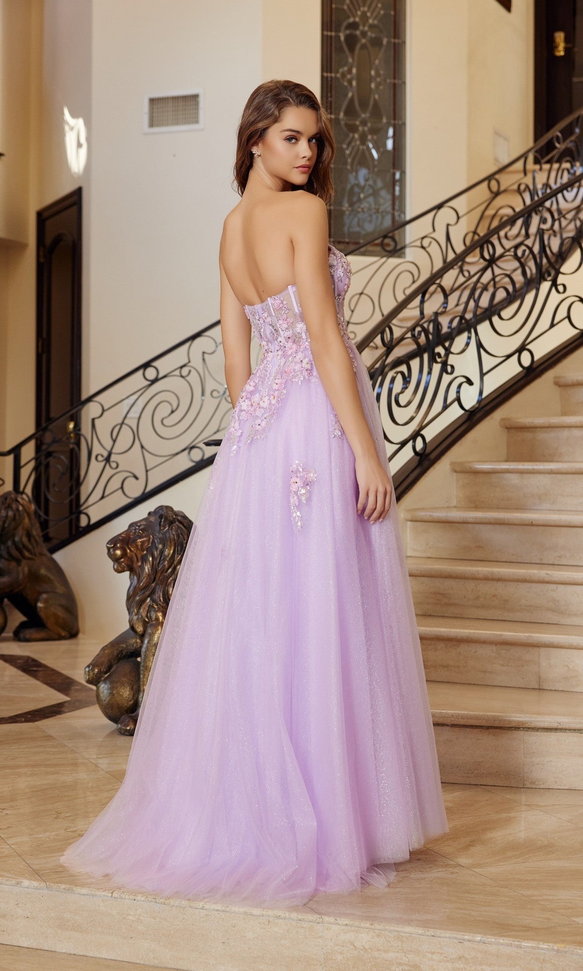 Wearing the Nox Anabel Q1773 long prom dress, a woman in a strapless lavender gown with 3D floral appliques and a corset bodice stands on a staircase, looking over her shoulder.